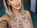 NicolSNaya has blonde hair, pink lipstick, and a patterned, see-through top showing her tits. She's tattooed with a black man's face on her arm.