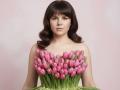 A portrait of Tilliana with dark, wavy hair and light skin. She stands against a pink background, holding a bouquet of vibrant pink tulips that covers her chest. The tulips are fresh, with green stems and leaves. Her expression is neutral, and she has soft makeup. The composition is centered, with the bouquet forming a horizontal line across her upper body. The image is clean and focused on the contrast between her hair and the pink tulips.