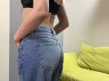 Stefanni's ass is prominent in blue jeans, with hands in pockets. She's wearing a black bra, and her back is slightly arched. A green pillow is in the background.