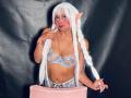 AilenRosse with white elf ears, long white braids, blue lace lingerie, red face makeup, holding braids, gold necklace, pink chair, black backdrop, tan skin, medium tits.