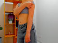Steisii with a slim, flat-stomach, wearing a gray crop top, blue high-waisted jeans, and a pink bracelet, covering her small tits with one arm.