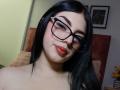Isabella Rossi, avec des lunettes noires, du rouge à lèvres, et des cheveux noirs lisses, souriant légèrement, montrant sa poitrine et son joli visage dans un selfie..