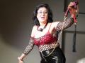 KarlaHartley with black hair, glasses, and red-black fishnet top showing her tits, holding a whip, wearing black latex pants, in a dominant, sexy pose.