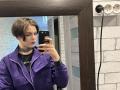 HanhFontillas with short brown hair, purple jacket, black shirt, choker, holding phone in mirror selfie, white brick wall, wooden mirror frame, black plugs, serious expression, looking at phone.