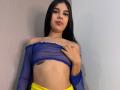 NaomyRosse with long black hair, blue sheer off-shoulder top, yellow pants, right hand on her chest, small tattoo under left breast, red lipstick, slight smile.