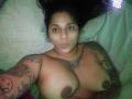 Chili, a topless woman with tan skin and dark hair, lies on a bed, showing off her large, round tits and tattoos, with a neutral expression, looking at the camera.