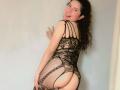EmilyCM with curly black hair, smiling, wearing a black lace fishnet dress, showing her ass, standing against a wall, making a peace sign with her right hand.