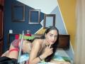 AfroditaHan with black hair, tan skin, small tits, wearing a white bra, red plaid skirt, and white thong, finger to lips, lying on bed, in a bedroom.