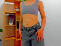 Steisii has a slim, flat-stomached body, small tits, wearing a grey sports bra and blue jeans with hands in pockets, standing next to a wooden shelf.
