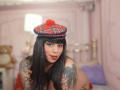 Chili has black hair, red lipstick, and a plaid hat with a red pom-pom. She's topless, showing tattoos on her arms, and has large, visible tits.