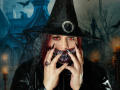 VanesaMoka with red eyes, black witch hat, and lace veil, has dark red hair, black nails, and a creepy, toothy grin, wearing a black jacket, in a spooky shot.