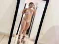LorensDouce standing in front of a mirror wearing pink lace lingerie, high heels, showing her tits, ass, and a tattoo on her side, taking a selfie with her phone.