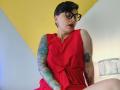 KatParadise with short black hair, glasses, red dress, tattoos on arms, sitting, looking down, red lipstick, yellow wall, white and yellow background, lamp on right.
