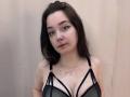 JuleeKasprzak with pale skin, dark brown hair, black strappy bra showing big tits, winged eyeliner, red lipstick, necklace, standing against a beige wall, looking at camera.