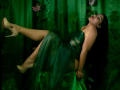 MariaRowe lying on green fabric, legs raised, wearing a green dress, gold high heels, dark hair, red lipstick. Green and black lighting, flowers in background. Sultry, seductive pose.