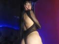Boorbuja with long black hair, wearing a black mesh bodysuit, exposing her back and butt, looking over her shoulder with a seductive gaze. Her ass is prominent.