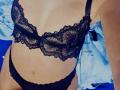 Der schwarze Spitzen-BH und die passenden schwarzen Unterhosen von YasminForLove, die ihre Brüste und ihre Vulva zeigen, mit ihrer gebräunten Haut und einem blauen Laken im Hintergrund..