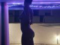 Silhouetted JuneLaFrenchie, naked, standing with arms behind back, showing curvy ass and small tits, under purple LED lights, in a dimly lit room.