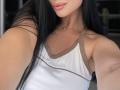 Olieri with long black hair, wearing a white tank top with brown trim, showing her tan skin, small necklace, and pink lips, in a selfie. Boobs visible, no ass.