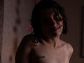 MaryCruzz, topless, with curly black hair, light skin, small tits, "Belletti" tattoo on right shoulder, dimly lit, slight smile, shadow on face, pink flowers in background.