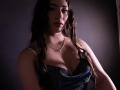 AnnFisher with long brown hair in a dark, sparkly dress showing her boobs, wearing a necklace, in dim lighting, looking at the camera with a serious expression.