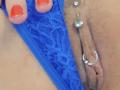 De blauwe kanten slip van FrenchShanna is naar de zijkant getrokken, waardoor haar geperforeerde vulva zichtbaar is, met een ring en een kralen. Haar oranje gelakte nagels zijn aan de linkerkant te zien..