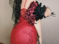 Niferttiti in a red, sheer lace dress with floral embroidery, holding black roses, showing her big ass and back, with long black hair, a tattoo, and a ring.