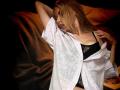 xMilagrosxss with blonde hair, eyes closed, wearing an open white shirt, black lace bra, hand on hip, other hand in hair, sitting on brown blanket.