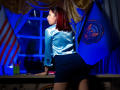 StephanieSun with red hair, shiny blue blouse, and black skirt, leaning on a desk, showing her ass, with flags and a window in the background, blue light.