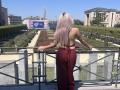EliseMonet with long platinum blonde hair, back to camera, wearing a red, high-waisted, cut-out dress, standing on a balcony, arms resting on a railing, looking at a garden.
