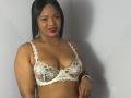 AliceBooty with medium brown skin, black hair, red lipstick, wearing a white lace bra with pink flower embroidery, showing her tits. She's smiling slightly, looking at the camera.