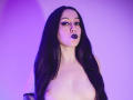 KendraSimons with long black hair, purple eyeliner, dark lipstick, and exposed bare tits, standing against a purple-lit background. Her nipples are visible, and she's looking at the camera.