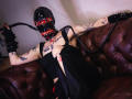 AmySpider with tattoos, wearing a black latex mask with red eyes, black dress, and gloves, holding a whip, sitting on a brown leather couch. Her pussy is covered.
