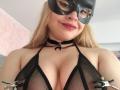 Melani Becker met blond haar, een zwart masker, rode lippenstift, een zwarte kanten BH, metalen klemmen op haar borsten, een zwarte choker met ketting, lachend, met haar decolleté zichtbaar, op een selfie..