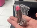 LillyMayer's hand holds a clear-wrapped chocolate dildo with a pink head, positioned vertically, showing the penis shape and texture.
