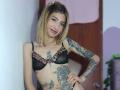 Kaiix with blonde hair, black bra with leopard print trim, and tattoos, smiling, hand on hip, showing off her tits and tattoos, pink hairclip.