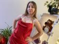 Aminas in a red satin dress, standing with hands on hips, showing small tits and a colorful tattoo on her left shoulder. She has light skin and brown hair.