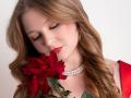AkilahGhil with long blonde hair, red lipstick, and pearl necklace, gently holding a red rose to her nose. Her eyes are closed, looking peaceful.