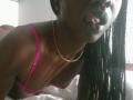 BettyBoopchi with dark skin, long braids, sticking out her tongue, wearing pink bra, gold necklace, leaning forward on bed, showing her tits and collarbone.