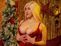 MikasaRosee with blonde hair, red dress, shiny boobs, pearl necklace, hands on chest, Christmas tree, gold star decorations, orange and gold background, looking to the side, smiling.