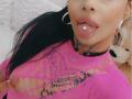 DenyChantal with black hair, pink lace top, tongue out, tongue piercing, hoop earrings, black choker, tattooed neck, and pink lipstick, showing her large tits and tattoos.