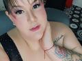 GiiannaMorrys with pink eyeshadow, glossy pink lips, black polka-dot dress, tattoo on right arm, red bracelet, and hand on neck, short brown hair, looking at camera.