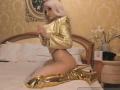 Mademoiselle Bella, con i capelli biondo platino, una maglia dorata, uno slip nero e stivali alti fino alla coscia dorati, è inginocchiata sul letto, mostrando il sedere, in una stanza con decorazioni dorate..
