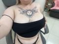 DiLaria with sun tattoo on chest, black strapless top, showing her big boobs, black thong, sitting in chair, blonde hair, white skin, office setting, visible chair and shelf.
