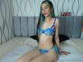 AbbyHillsI with blue lace bra and panties, sitting on bed, showing small tits, blue floral pattern, long green and brown hair, light skin, smiling, wristwatch, green nail polish.