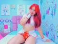 MiaLeroy with red hair, white crop top, red thong, and white mini skirt, sitting on bed, showing her pussy, cute smile, hand near mouth, dreamcatchers on wall.
