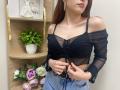 Rioria with small tits in a black sheer off-shoulder top, pulling it down slightly, wearing blue jeans, gold earrings, and pink lipstick.