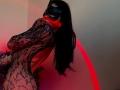 Tsukerberg in a black lace bodysuit, mask covering eyes, long black hair, red lighting, back to camera, hand over mouth, showing her ass, looking over shoulder.