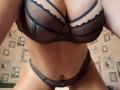 Dinastiia's big, dark mesh tits in a black bra with lace, showing her tan, curvy belly and matching black panties with a zipper, leaning forward.