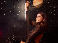 LucieMilner, with sparkling face makeup, wearing a sheer black top and glossy black pants, gripping a pole, smiling, her tits subtly visible through the mesh.